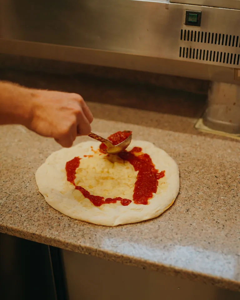 Eine Hand streicht Tomatensoße mit der Kelle auf einen Pizzaboden