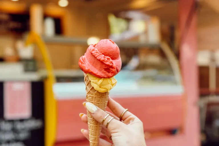 Eine Hand hält ein Hörnchen mit zwei Kugeln Eis vor dem altrosa Eiswagen