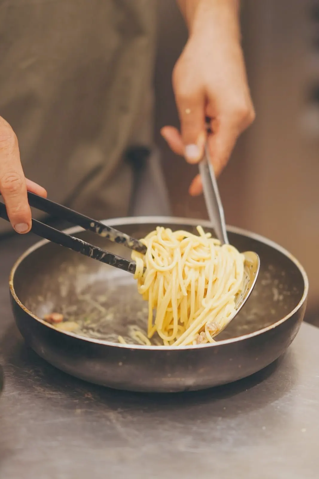 Zwei Hände heben Spaghetti mit einer Zange aus der Pfanne, in der Küche des Il Colosseo